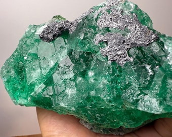 May include: A raw, uncut fluorite mineral specimen. The fluorite is a vibrant green color with a crystalline structure. A dark, metallic-looking mineral is embedded in the fluorite.