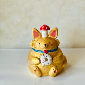 May include: A golden ceramic figurine of a fox wearing a blue and white collar with a red heart charm. The fox is holding a donut with sprinkles and has a red and white mushroom on its head.