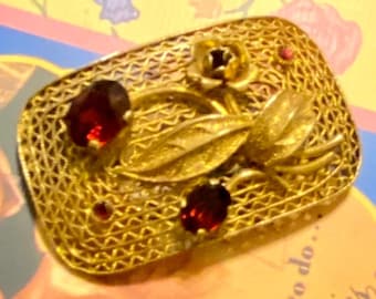 Op de afbeelding: Een gouden filigraan broche met rode glazen stenen. De broche heeft een bloemmotief met bladeren en een enkele roos.