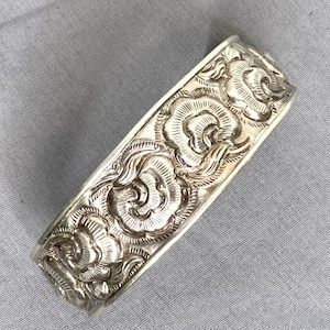May include: A silver-colored cuff bracelet with an ornate floral design. The bracelet features a detailed pattern of swirling flowers and leaves, creating a textured surface. The metal has a polished finish, reflecting light.