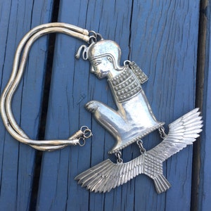 May include: A silver necklace with a pendant featuring a stylised figure of a person with wings. The pendant is detailed with textured patterns and the figure is in a kneeling position.