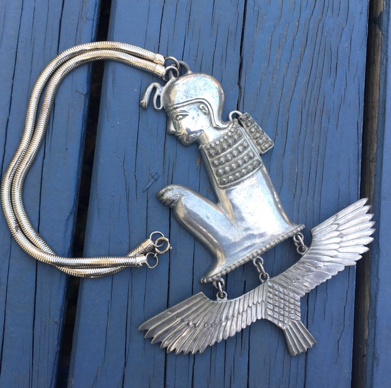 May include: A silver necklace with a pendant featuring a stylised figure of a person with wings. The pendant is detailed with textured patterns and the figure is in a kneeling position.