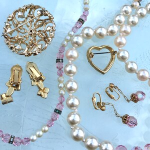 May include: A set of vintage jewelry featuring a large gold brooch with a floral design, a pair of gold earrings with a floral design, a heart-shaped gold brooch, and two strands of pearls with pink crystal beads.