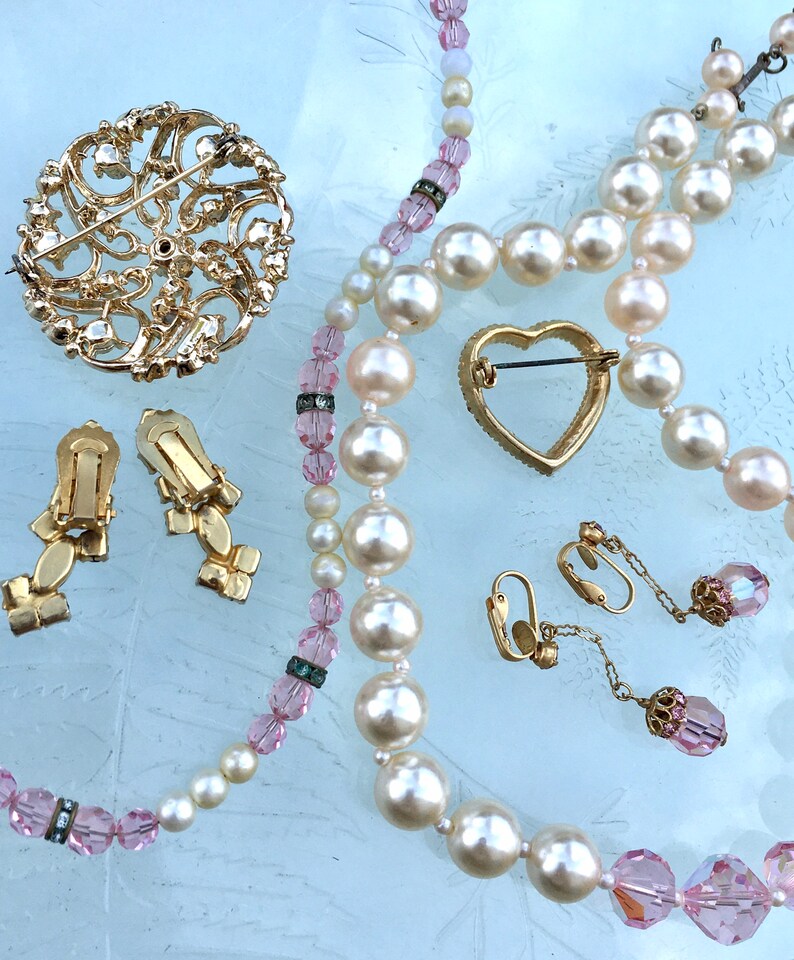 May include: A set of vintage jewelry featuring a large gold brooch with a floral design, a pair of gold earrings with a floral design, a heart-shaped gold brooch, and two strands of pearls with pink crystal beads.