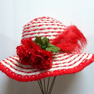 Puede incluir: Un sombrero de sol a rayas blancas y rojas con una pluma roja y una flor de clavel roja.