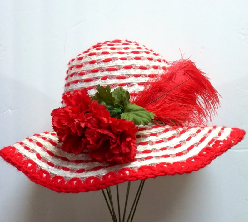 Puede incluir: Un sombrero de sol a rayas blancas y rojas con una pluma roja y una flor de clavel roja.