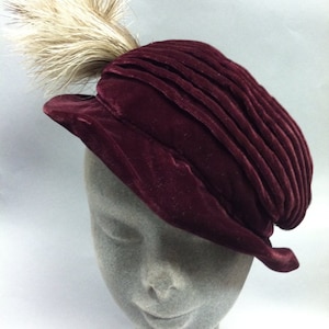 May include: A burgundy velvet hat with a pleated design and a white feather accent.
