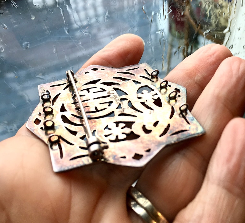 May include: A silver-toned, star-shaped brooch with intricate cut-out designs. The brooch has a pin and catch closure on the back.