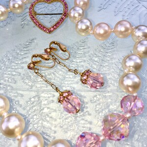 May include: A pearl necklace with a pink heart-shaped brooch and pink crystal earrings. The necklace is made of white pearls and the earrings are made of gold and pink crystals.