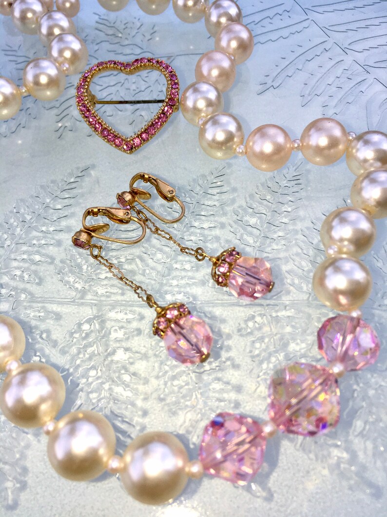May include: A pearl necklace with a pink heart-shaped brooch and pink crystal earrings. The necklace is made of white pearls and the earrings are made of gold and pink crystals.