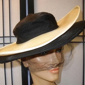 May include: A black and cream wide-brimmed hat with a black veil. The hat has a large, floppy brim and a crown that is slightly tilted.