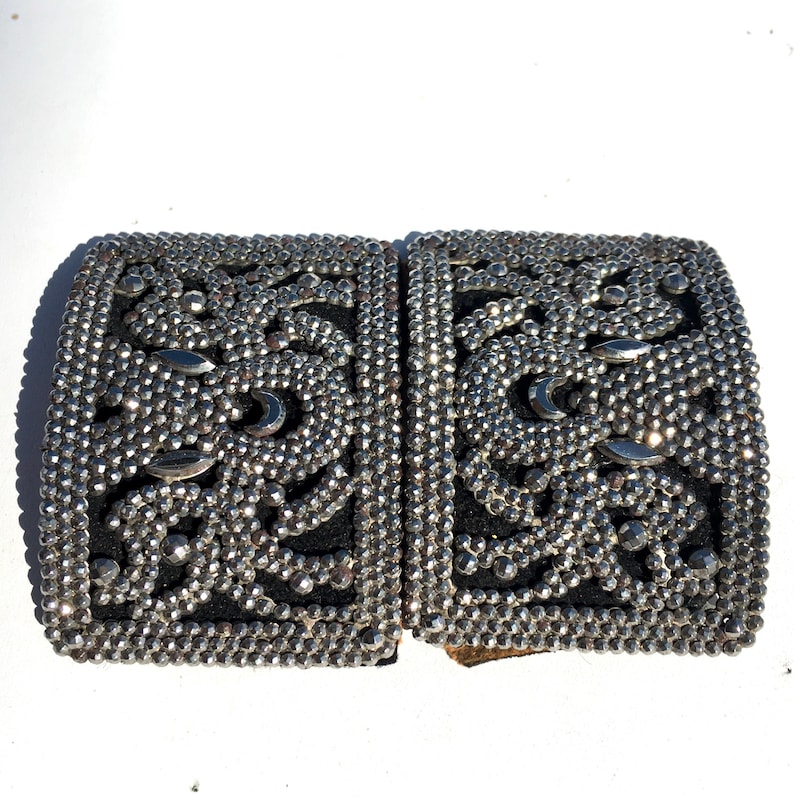 May include: A pair of silver-toned metal belt buckles with a decorative design of small round studs and a geometric pattern. The buckles are joined together.