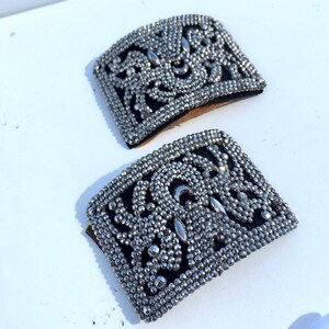 May include: Two silver-toned metal embellishments with a black fabric backing. The embellishments are rectangular and feature a decorative design of small metal beads.