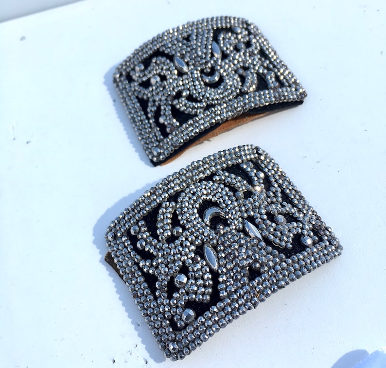 May include: Two silver-toned metal embellishments with a black fabric backing. The embellishments are rectangular and feature a decorative design of small metal beads.