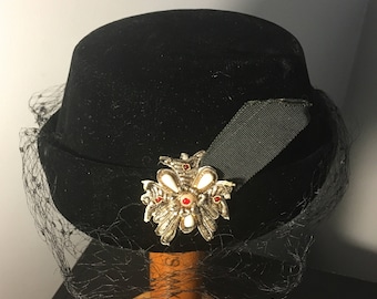 May include: A black velvet hat with a black net veil and a jeweled brooch. The brooch is silver with red and white stones.