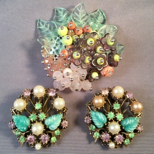 Può includere: Un set di gioielli vintage composto da una spilla e orecchini. La spilla è un gruppo di fiori rosa, verdi e viola con foglie. Gli orecchini sono di design abbinato con foglie verdi e perle rosa e bianche.