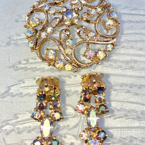 May include: A gold tone brooch and earrings set with iridescent rhinestones. The brooch is a circular design with a filigree pattern. The earrings are dangle style with a teardrop shaped rhinestone.