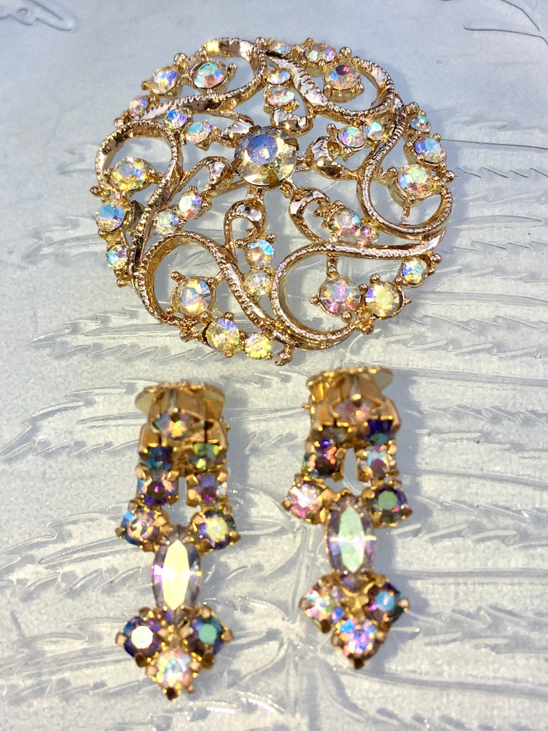 May include: A gold tone brooch and earrings set with iridescent rhinestones. The brooch is a circular design with a filigree pattern. The earrings are dangle style with a teardrop shaped rhinestone.