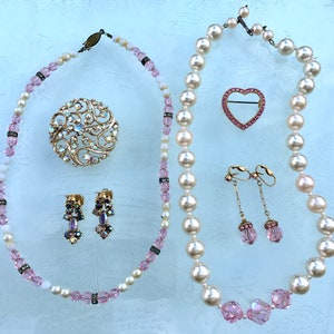 May include: A set of vintage jewelry featuring a pearl necklace, a pink heart brooch, a floral brooch, and a pair of dangle earrings with pink stones.