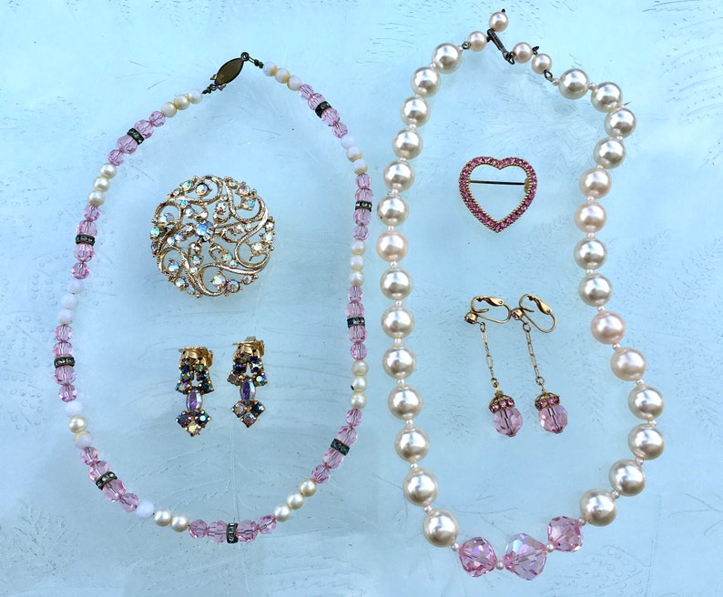 May include: A set of vintage jewelry featuring a pearl necklace, a pink heart brooch, a floral brooch, and a pair of dangle earrings with pink stones.