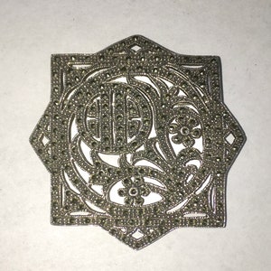 May include: A silver brooch with a geometric design, featuring a floral motif and a circle in the center. The brooch is covered in small, sparkling stones.