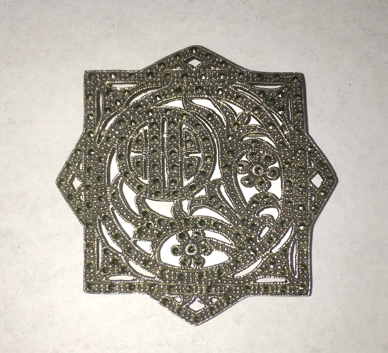 May include: A silver brooch with a geometric design, featuring a floral motif and a circle in the center. The brooch is covered in small, sparkling stones.