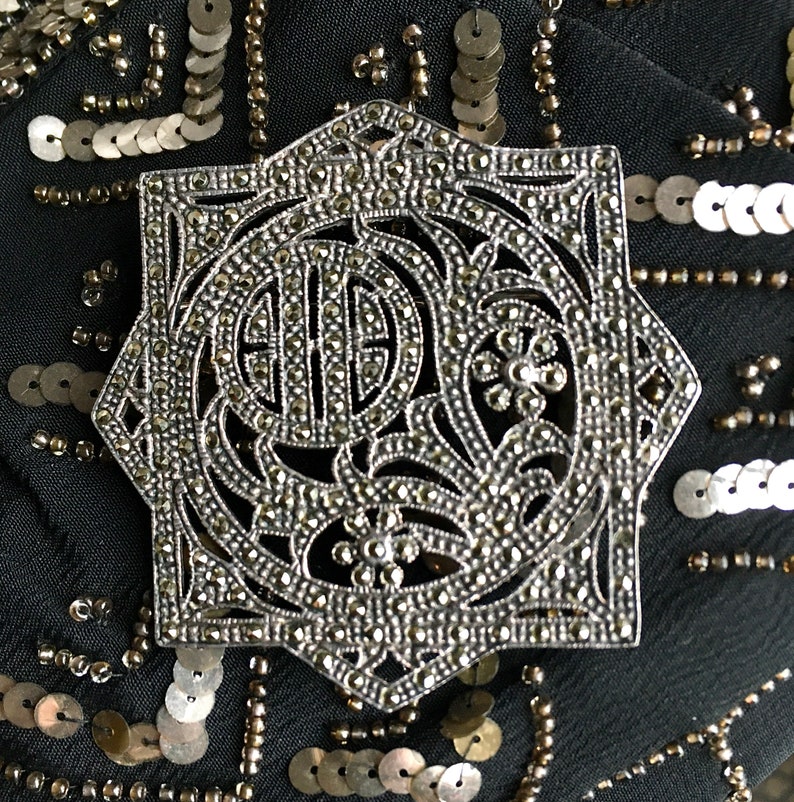 May include: A silver-toned, octagonal-shaped brooch with a detailed, intricate design. The brooch is covered in small, sparkling stones.