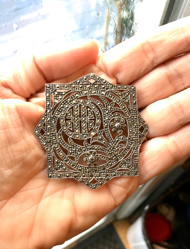 May include: A silver and marcasite brooch with a geometric design and a floral motif. The brooch is in the shape of a star and has a filigree design.