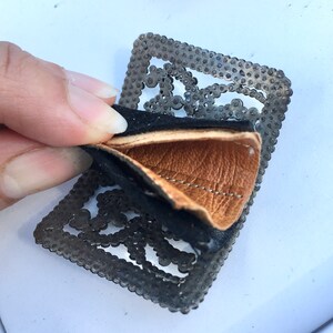 May include: A black and brown leather patch with a metal frame. The metal frame has a decorative design of small circles and squares.