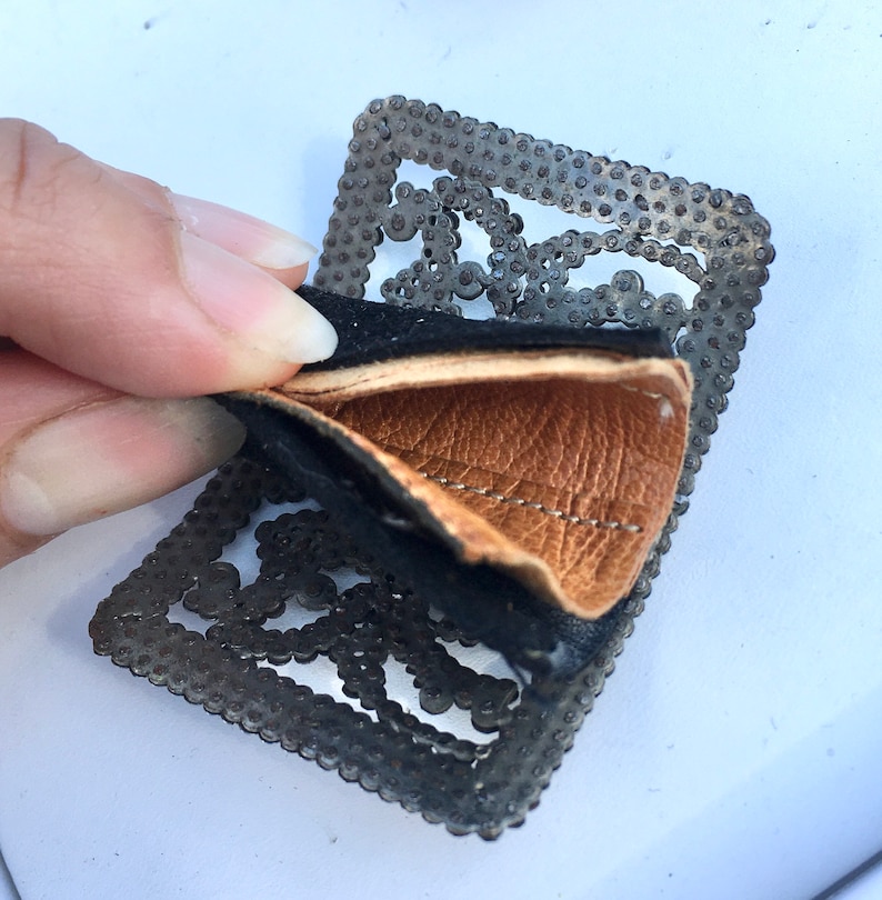 May include: A black and brown leather patch with a metal frame. The metal frame has a decorative design of small circles and squares.