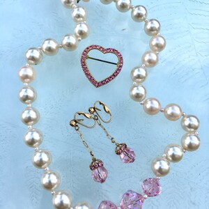 May include: A pearl necklace with a pink heart-shaped brooch and pink crystal earrings. The necklace is made of white pearls and the brooch and earrings are set with pink crystals.