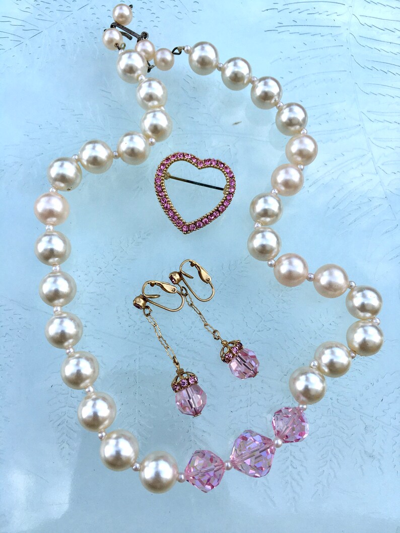 May include: A pearl necklace with a pink heart-shaped brooch and pink crystal earrings. The necklace is made of white pearls and the brooch and earrings are set with pink crystals.