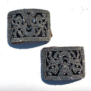 May include: Two silver metal buckles with a decorative design of small beads and a floral pattern. The buckles are rectangular and have a slightly worn look.
