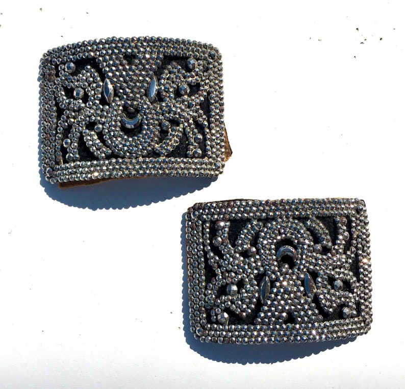 May include: Two silver metal buckles with a decorative design of small beads and a floral pattern. The buckles are rectangular and have a slightly worn look.