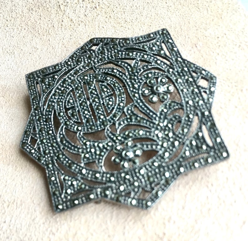 May include: A silver brooch with a star-shaped design, set with marcasite stones. The brooch is intricate and detailed, with a floral motif.