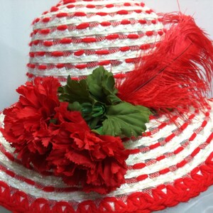 Puede incluir: Un sombrero de paja blanco y rojo a rayas con una pluma roja y claveles rojos.