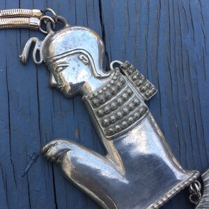 May include: Silver tone pendant necklace featuring a stylised figure of a person wearing a helmet and armour. The pendant is detailed with a textured surface and a chain.