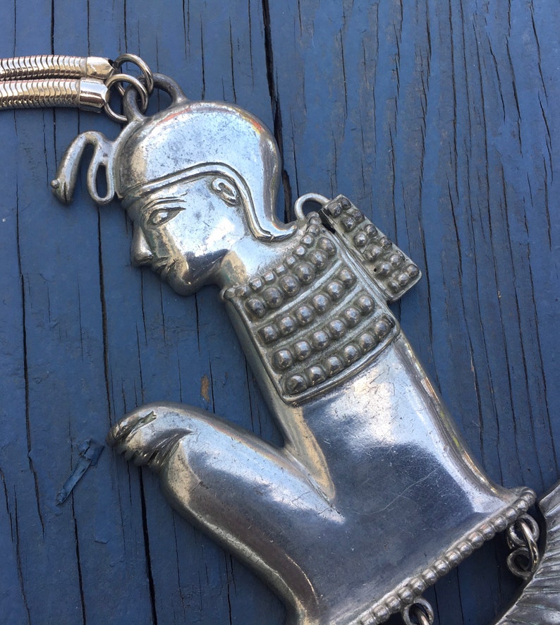 May include: Silver tone pendant necklace featuring a stylised figure of a person wearing a helmet and armour. The pendant is detailed with a textured surface and a chain.