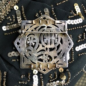 May include: Silver tone brooch with an intricate design featuring a circle with a Chinese character in the center. The brooch has a pin and catch closure.