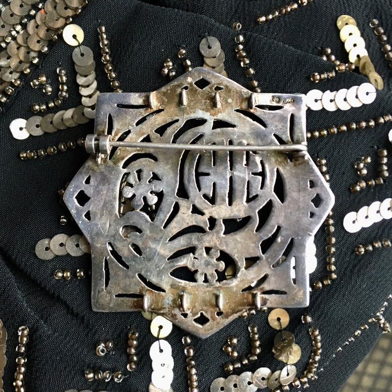 May include: Silver tone brooch with an intricate design featuring a circle with a Chinese character in the center. The brooch has a pin and catch closure.