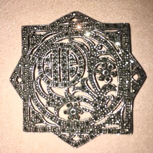 May include: A silver and black star-shaped brooch with a floral design and a central circle. The brooch is made of metal and is covered in small black stones.