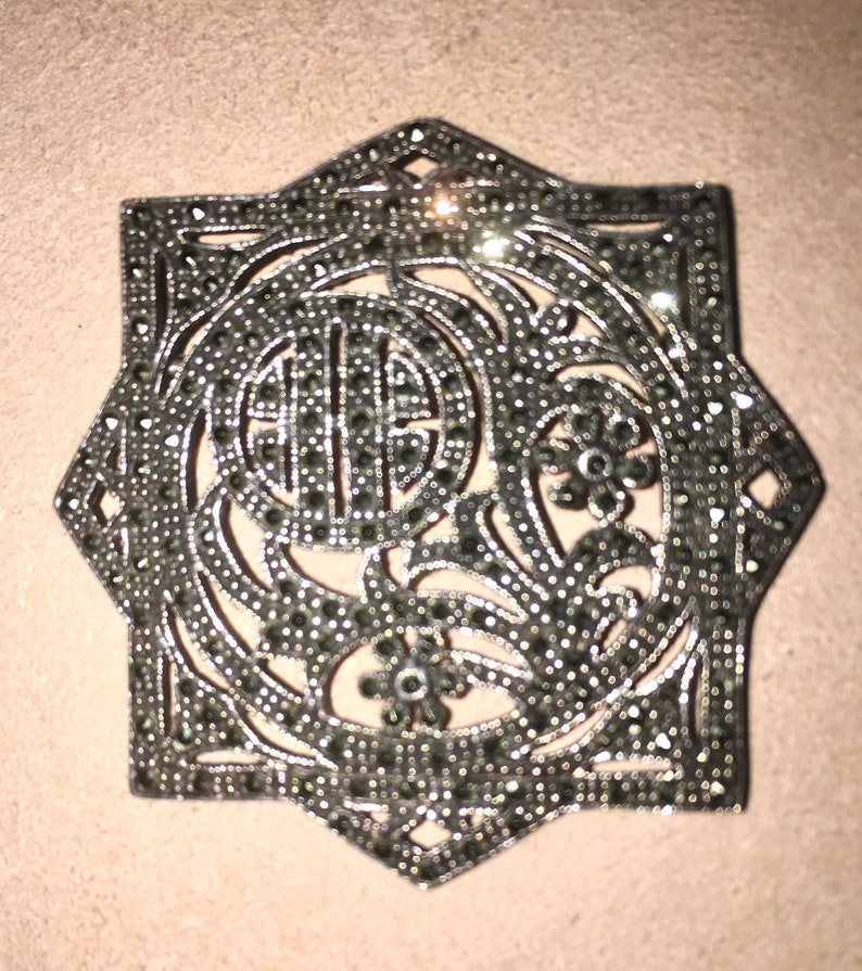 May include: A silver and black star-shaped brooch with a floral design and a central circle. The brooch is made of metal and is covered in small black stones.
