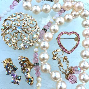 May include: A set of vintage jewelry featuring a pearl necklace, a heart-shaped brooch with pink rhinestones, a large brooch with iridescent rhinestones, and a pair of clip-on earrings with iridescent rhinestones.