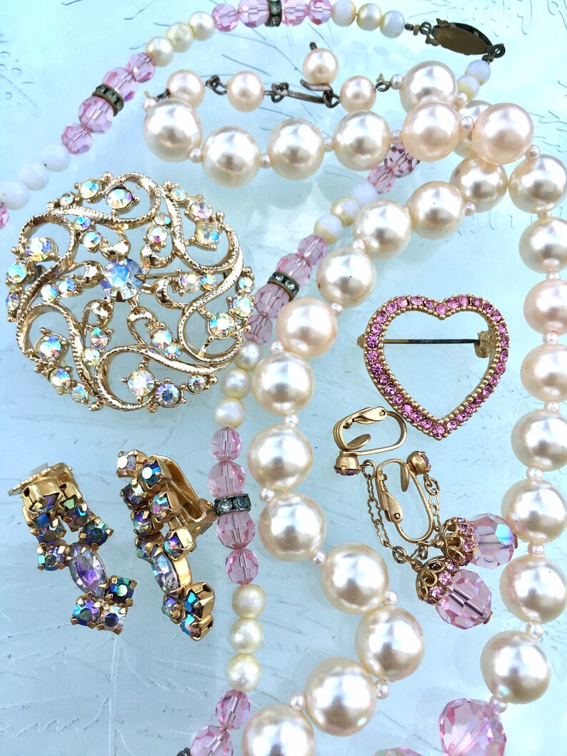 May include: A set of vintage jewelry featuring a pearl necklace, a heart-shaped brooch with pink rhinestones, a large brooch with iridescent rhinestones, and a pair of clip-on earrings with iridescent rhinestones.