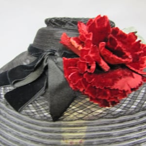 May include: A black net hat with a large red velvet flower accent. The hat has a wide brim and a black velvet ribbon tied in a bow.