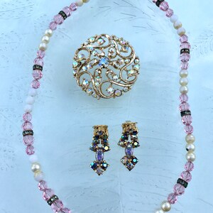 May include: A vintage jewelry set featuring a pink and white beaded necklace, a large iridescent brooch with a floral design, and a pair of dangling earrings with iridescent stones.