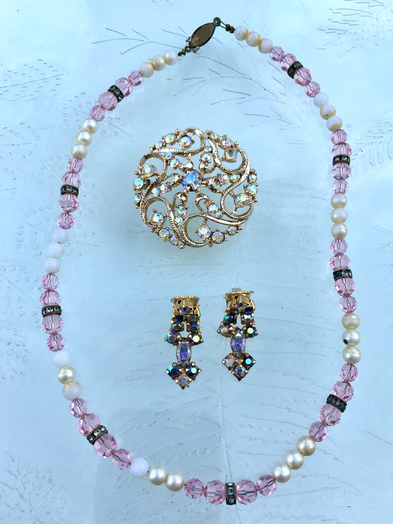 May include: A vintage jewelry set featuring a pink and white beaded necklace, a large iridescent brooch with a floral design, and a pair of dangling earrings with iridescent stones.