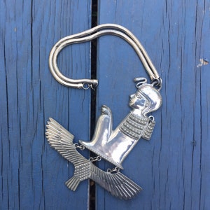 May include: Silver necklace with a pendant featuring a stylised figure with wings. The figure is wearing a crown and has a detailed design.