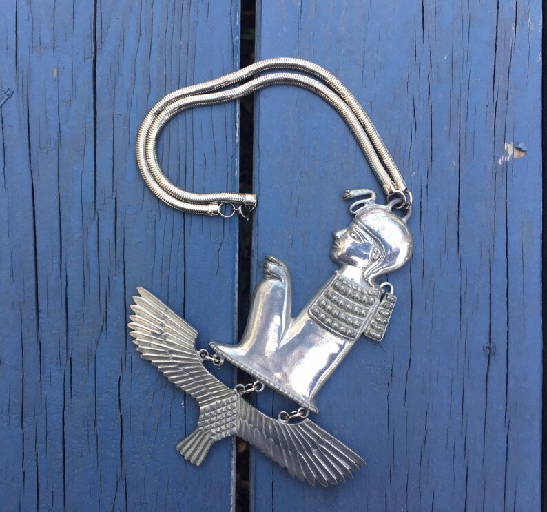 May include: Silver necklace with a pendant featuring a stylised figure with wings. The figure is wearing a crown and has a detailed design.