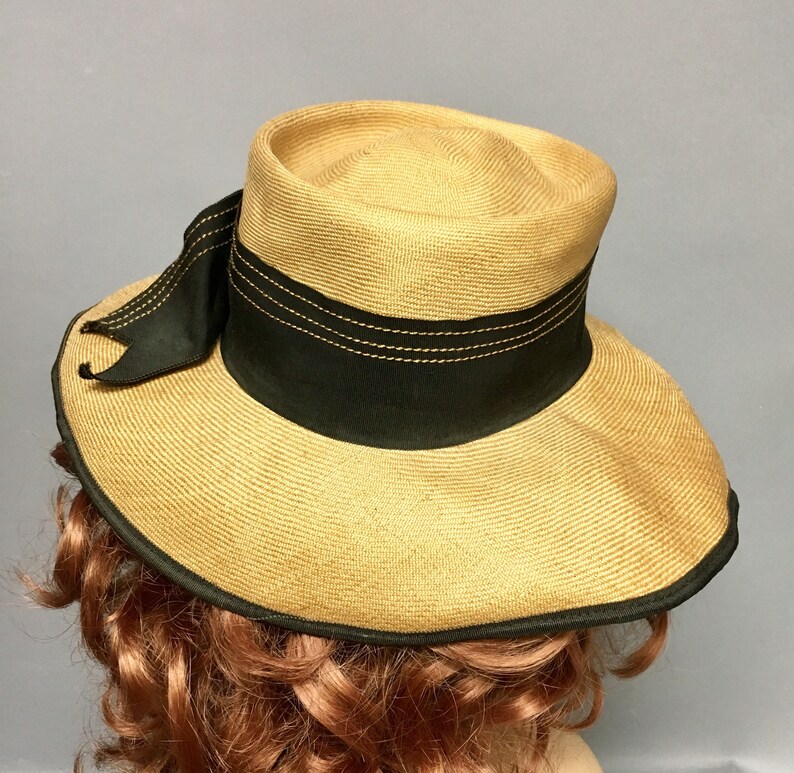 May include: A straw hat with a wide brim and a black ribbon around the crown. The hat has a black ribbon with a bow on the side.